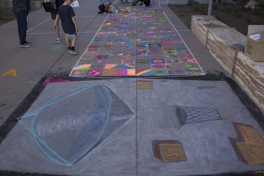This photograph shows the full-size Psyche spacecraft outline drawn in sidewalk chalk. The bus (body) was completed by the artist, Brianna, but individual squares on the solar panels were filled in by children and visitors with any space image they chose.