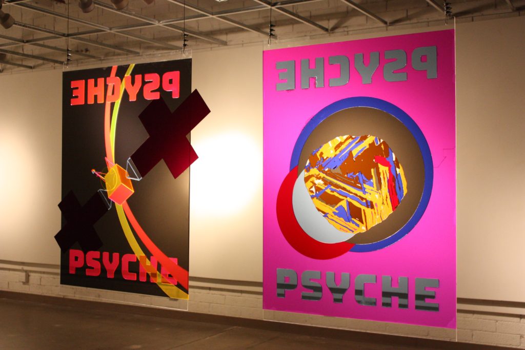 Two posters made from brightly-colored transparent acrylic cut and fit together; one shows the Psyche spacecraft, the other the asteroid. A colorful image is cast behind each poster when lit from one side.
