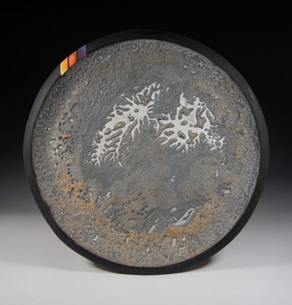 This image shows a circular ceramic platter. The center is grey with swirls and bubbles. The rim is black with three lines in colors of yellows, oranges, and purples.