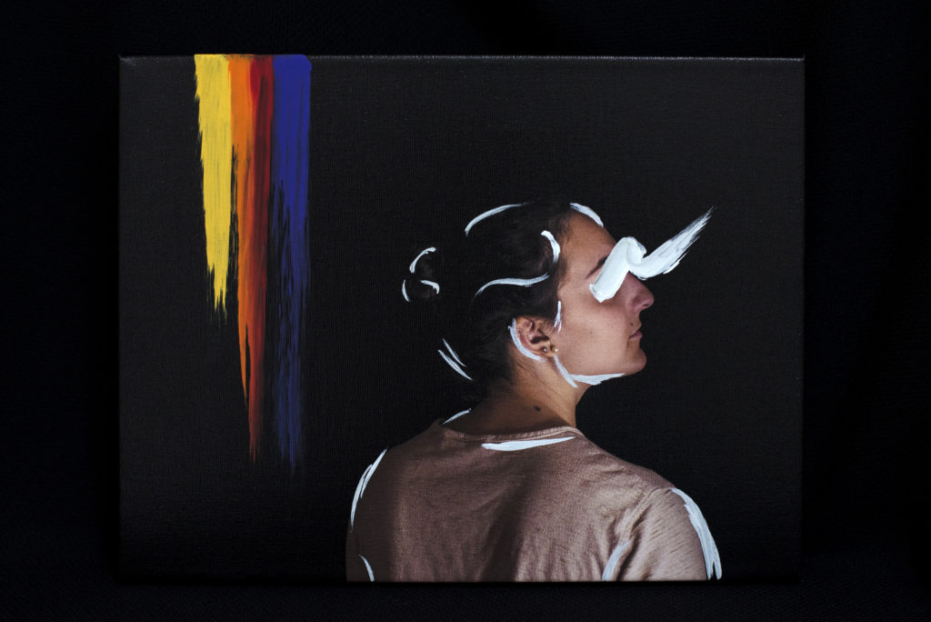 This is a photograph of a woman looking outwards and upwards. The background is gray and black and there are painted stripes of yellow, orange, red, and purple along one side. The woman's outline is accentuated and her eyes are obscured with white paint.