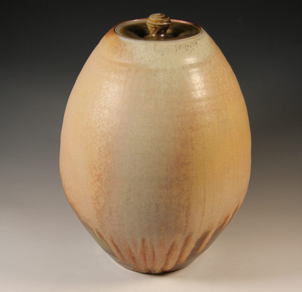 This image shows an oval ceramic vessel with a lid. The glaze is a mix of tans, dark reds, and browns with streaks that are prominent near the base. The lid has a textured knob on top.