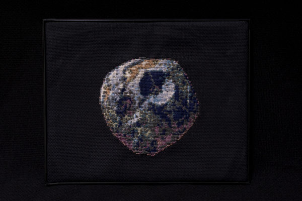 This image shows a cross stitch version of the artist's rendition of the Psyche asteroid on a black background. It shows the possible cratering and detail of the surface.
