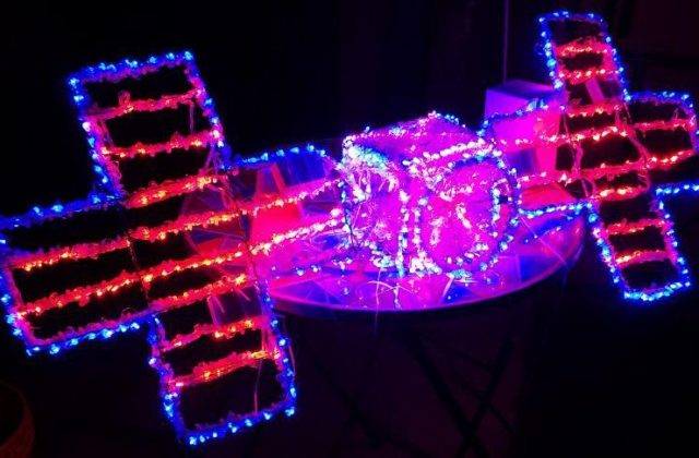 This image shows a sculpture made out of wire and LED holiday lights (pinks, reds, and blues) in the shape of the Psyche spacecraft.