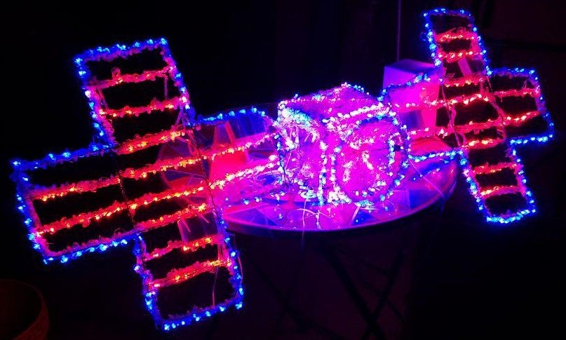 This image shows a sculpture made out of wire and LED holiday lights (pinks, reds, and blues) in the shape of the Psyche spacecraft.