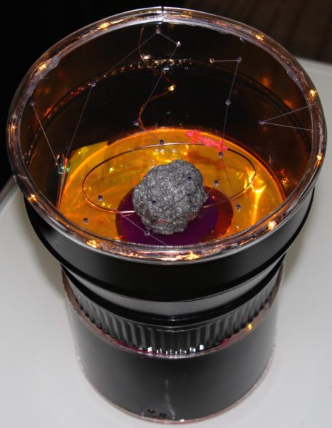 This image shows a diorama representing the end of a telescope. Inside it is the Psyche asteroid made of grey metal being orbited by a small pink acrylic Psyche spacecraft on a copper wire. The lens of the telescope has constellations etched into it its surface.