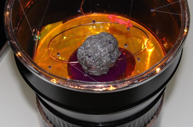 This image shows a diorama representing the end of a telescope. Inside it is the Psyche asteroid made of grey metal being orbited by a small pink acrylic Psyche spacecraft on a copper wire. The lens of the telescope has constellations etched into it its surface.