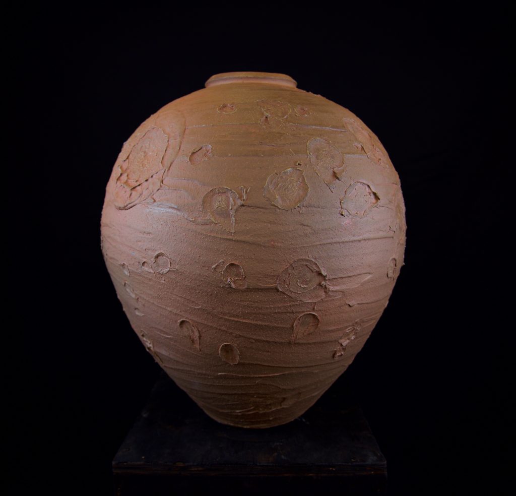 This image shows a large ceramic vessel, reddish in color and covered in rough craters and streaks.
