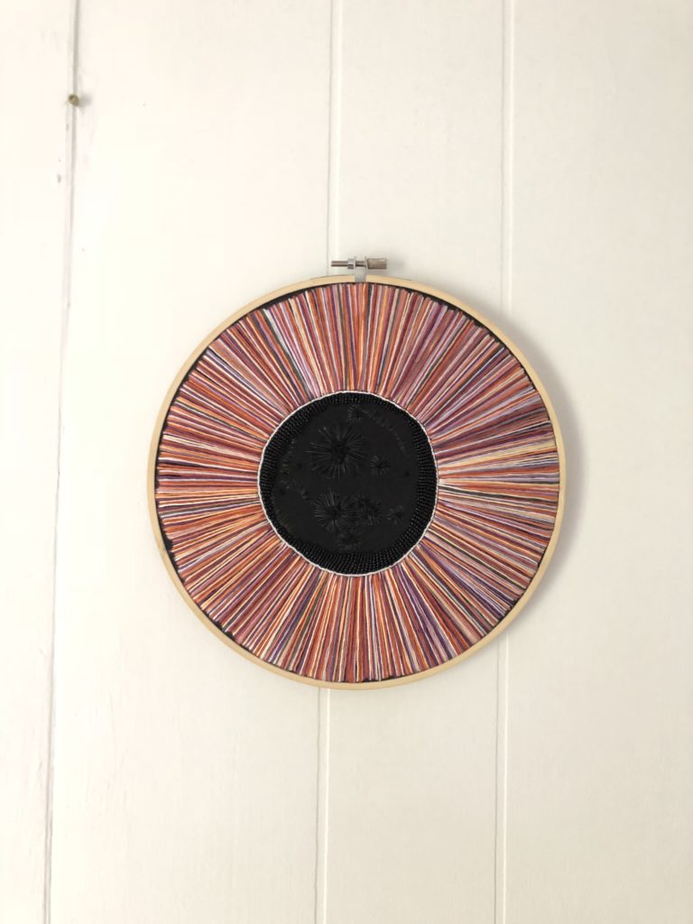 A 9” wooden hoop with Psyche’s silhouette outlined in black beading, surrounded by rays of purple and orange stretching outward to fill the hoop.