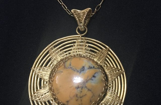 Circular pendant made from orange dendritic opal wrapped with brass wire, in a pattern shaped like the sun.