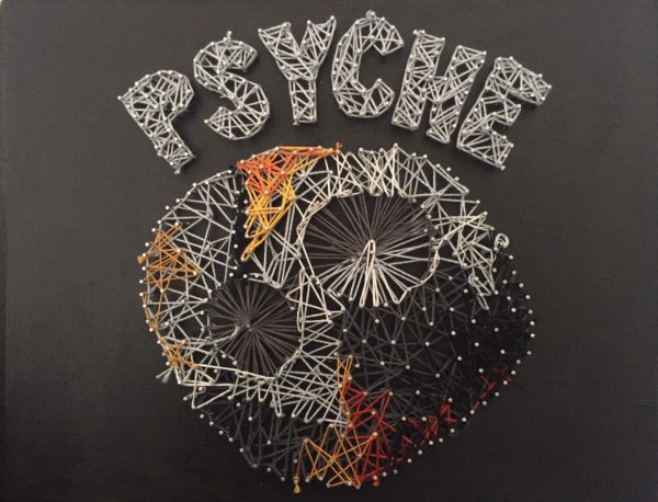 This piece features Psyche against a black wooden background. The shape of Psyche is composed of many tiny nails, with string wrapped around them. In this jumbled string, the two biggest craters stand out. They keep their signature circular shape. The asteroid has tints of orange and yellow at the bottom of it and on top, signifying metal. On top of the asteroid, also in string and nails, is the word PSYCHE in all caps and grey.