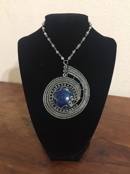 a necklace that has a blue central stone to represent the Earth and three thick silver wires wrapping around the stone similar to the Psyche spacecraft's flight path.