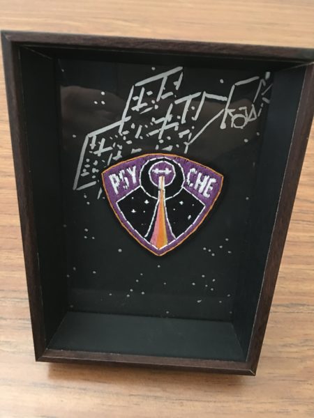 An embroidered rounded triangle, purple with a golden outline. Inside is a rainbow trail leading upwards towards a small image of the orbiter, two crosses with a square in the middle. The orbiter is surrounded by two circles. On either side of the circles are P S Y and C H E in white.