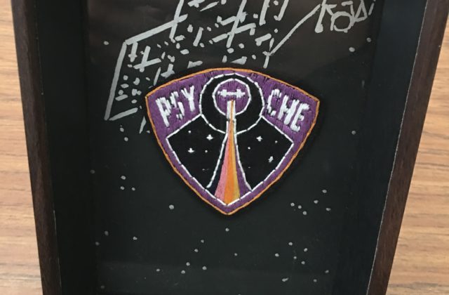 An embroidered rounded triangle, purple with a golden outline. Inside is a rainbow trail leading upwards towards a small image of the orbiter, two crosses with a square in the middle. The orbiter is surrounded by two circles. On either side of the circles are P S Y and C H E in white.