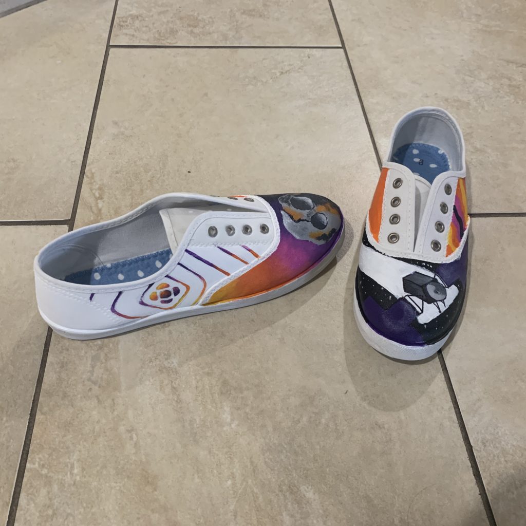 On the left shoe, at the toe area, there is a semi realistic painting of the Psyche asteroid. The background is a gradient from yellow to purple. On the outside of the left shoe is the logo for Psyche Inspired. The diamond shaped lines that have a similar gradient is extended to cover the whole outside of the shoe. The inside side of the left shoe is a vibrant orange. On the right shoe, at the toe area, is the spacecraft. It is slightly tilted, cutting off by the edges. The background is black and starry. The outside side of the right shoe is meant to be a "groovy" pattern. There are wavy stripes alternating between the signature yellow, orange, pink, and purple found in the logo. The inside side of the right shoe is also a vibrant orange. Keeping both insides a vibrant orange brings the shoe together and makes them seem more like a pair of shoes rather than two completely different shoes with different designs.