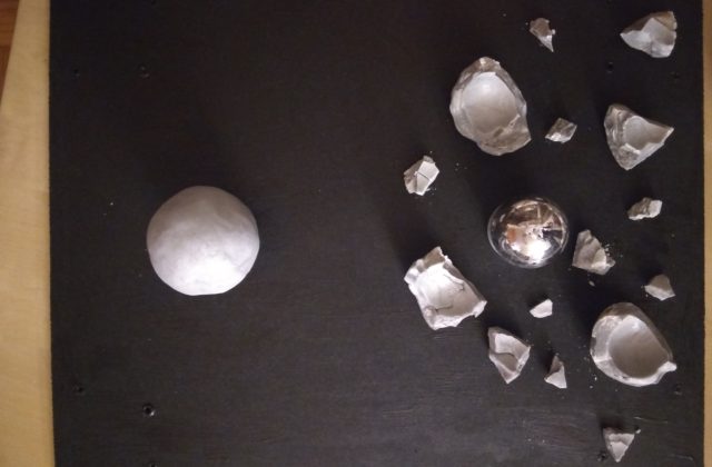 The video shows a gray ball being smashed into fragments by a rock, revealing a silver ball inside. The photo shows the fragments and silver ball arranged on a black board, with another gray ball nearby.