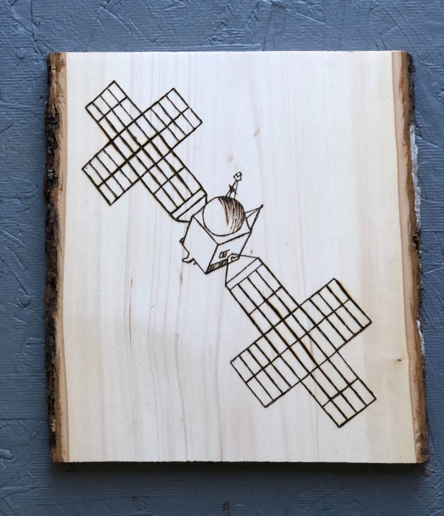 This image shows a vertical slice of wood with an image of the Psyche spacecraft made using woodburning.