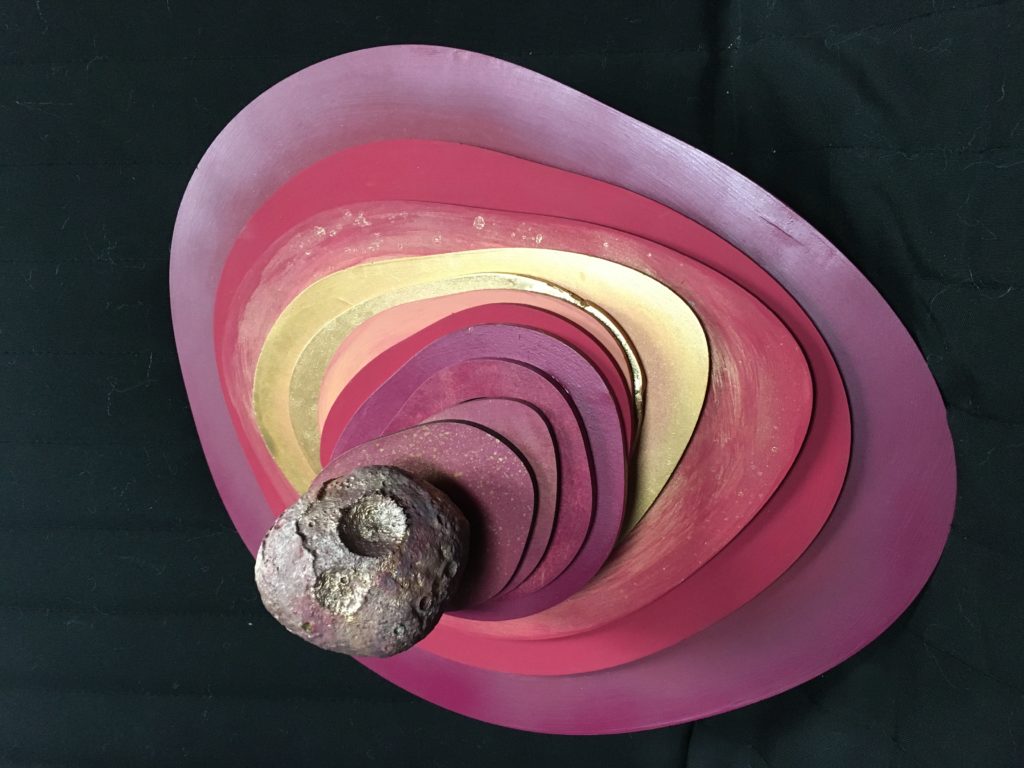 Foot-by-foot concentric organic shapes are stacked and spiraling towards the viewer in pinks, oranges and golds leading towards the gray, 3D printed Psyche Asteroid. Dry brushed pinks and gold follow the contour lines of the 3D print.