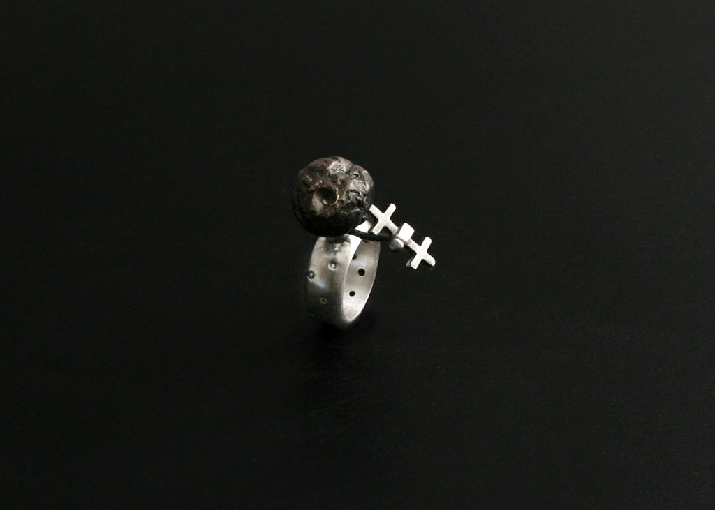 This image shows a sterling silver kinetic ring with a darkened bronze asteroid as the centerpiece. A tiny spacecraft placed next to the asteroid is mobile. The small spacecraft can be pushed around the asteroid to simulate orbit.
