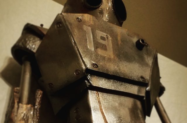 A robot stands 6ft tall with the number 19 displayed on its chest. There is rust coming from the edges of the plating. There are dents and scratches covering the robot’s head, arms, and chest. It seems to be looking at something in the distance with dark, black lenses–one of them larger than the other. The background is white, bringing a dynamic contrast to the dark bronze metallic glow of the robot’s plating.