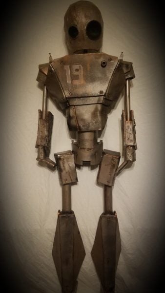 A robot stands 6ft tall with the number 19 displayed on its chest. There is rust coming from the edges of the plating. There are dents and scratches covering the robot’s head, arms, and chest. It seems to be looking at something in the distance with dark, black lenses–one of them larger than the other. The background is white, bringing a dynamic contrast to the dark bronze metallic glow of the robot’s plating.