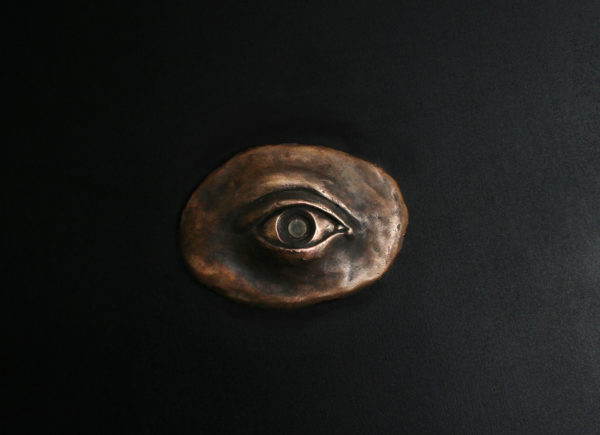 An oval-shaped art piece cast in bronze in the shape of an eye with a glass pupil. The pupil is a magnification lens through which one can view an image of Psyche in human form looking up at a spacecraft traveling toward her.