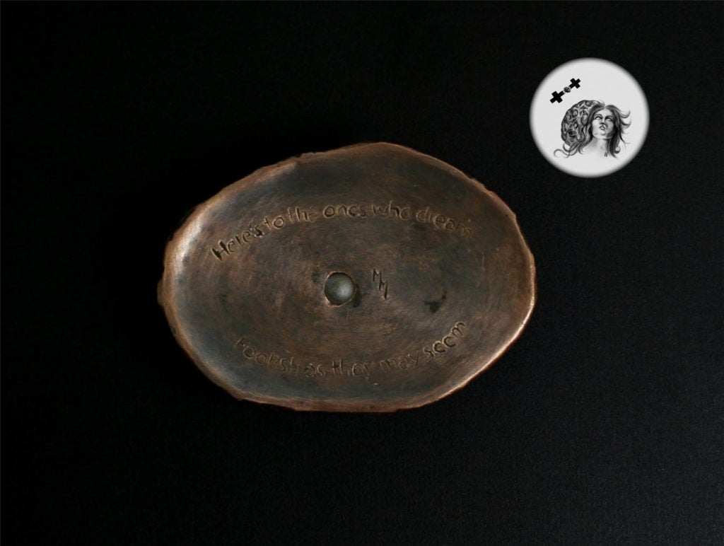 An oval-shaped art piece cast in bronze in the shape of an eye with a glass pupil. The pupil is a magnification lens through which one can view an image of Psyche in human form looking up at a spacecraft traveling toward her.