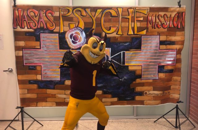 The ASU mascot standing in front of the NASA Psyche Mission backdrop created by a Psyche Inspired intern.