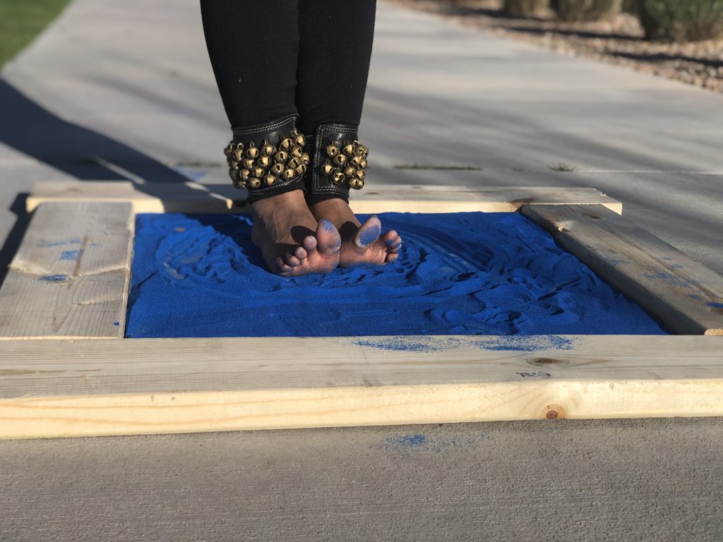 A dancer's feet are posed and tracing an image in bright blue sand poured into a framed flat surface. The traced image is a simplistic representation of the Psyche spacecraft orbiting the asteroid (16) Psyche