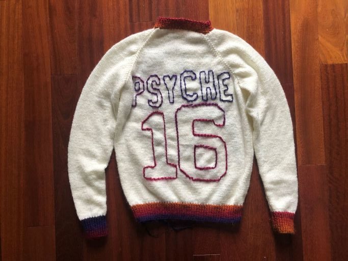 On the front, left side of a white sweater, there's a purple outline of the Psyche spacecraft with a small pink signal wave. On the back of the sweater is a large number 16 with the word “PSYCHE” spelled in all caps, placed like the design of a sports jersey. The collar, sleeve cuffs, lower band, letters, and numbers of the sweater are in an ombre yarn of purple, pink, red, and orange.