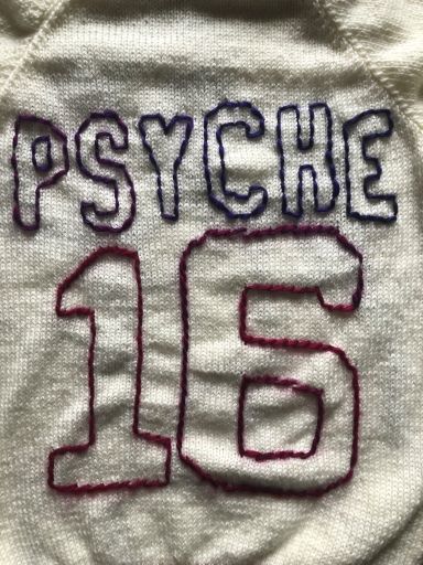 On the front, left side of a white sweater, there's a purple outline of the Psyche spacecraft with a small pink signal wave. On the back of the sweater is a large number 16 with the word “PSYCHE” spelled in all caps, placed like the design of a sports jersey. The collar, sleeve cuffs, lower band, letters, and numbers of the sweater are in an ombre yarn of purple, pink, red, and orange.