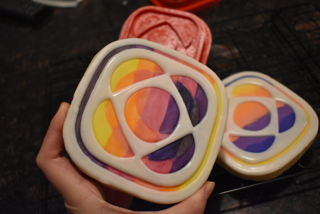 An asteroid-shaped cookie of the Psyche mission badge with black lettering that says, "Journey to a Metal World PSYCHE," is centered in the foreground. Mission badge and asteroid cookies of the same yellow, orange, pink, and purple gradient are in the background.