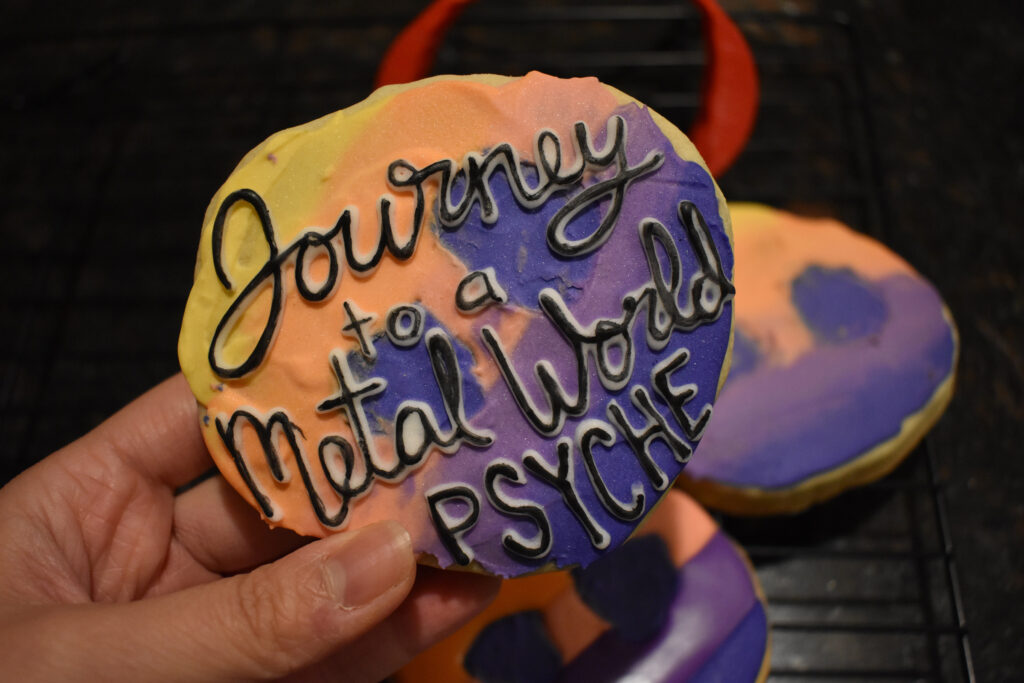 An asteroid-shaped cookie of the Psyche mission badge with black lettering that says, "Journey to a Metal World PSYCHE," is centered in the foreground. Mission badge and asteroid cookies of the same yellow, orange, pink, and purple gradient are in the background.