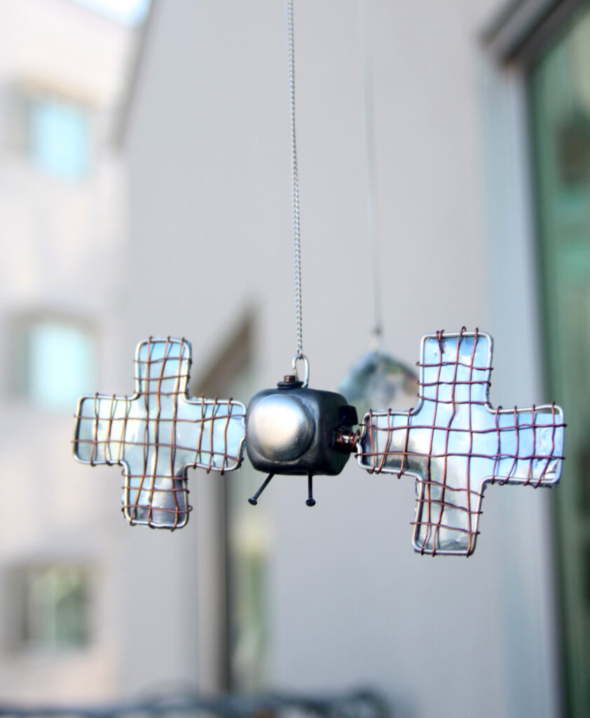 This is a hanging mobile made from wire, with clay pieces of Psyche and the satellite hanging by chains. They are painted in a shiny metallic paint that shimmers in the sun and are hanging with two sun catcher crystals.