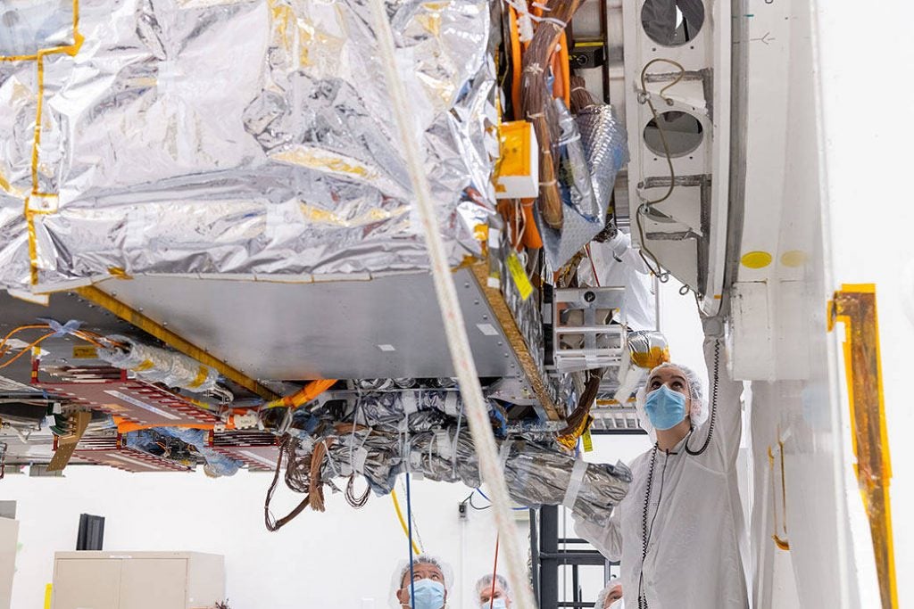 This image is a closeup underside view of the main body of the spacecraft (the bus) with a engineers nearby inspecting hardware.