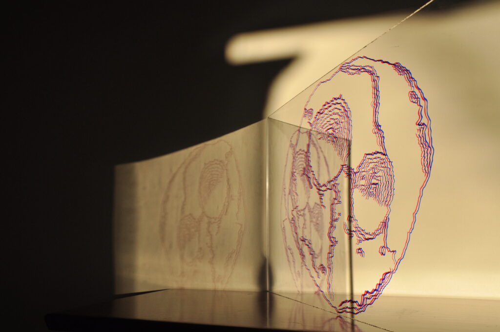 This piece is 24 inches by 13.5 inches. Red and black ink marks on 0.25 inch plexiglass were utilized to create lines that outline the shape of Psyche. There are two major craters and a series of rifts on the surface that appear pockmarked with small craters. As light shines through the plexiglass, red and blue shadows in the shape of Psyche are cast onto a white background.