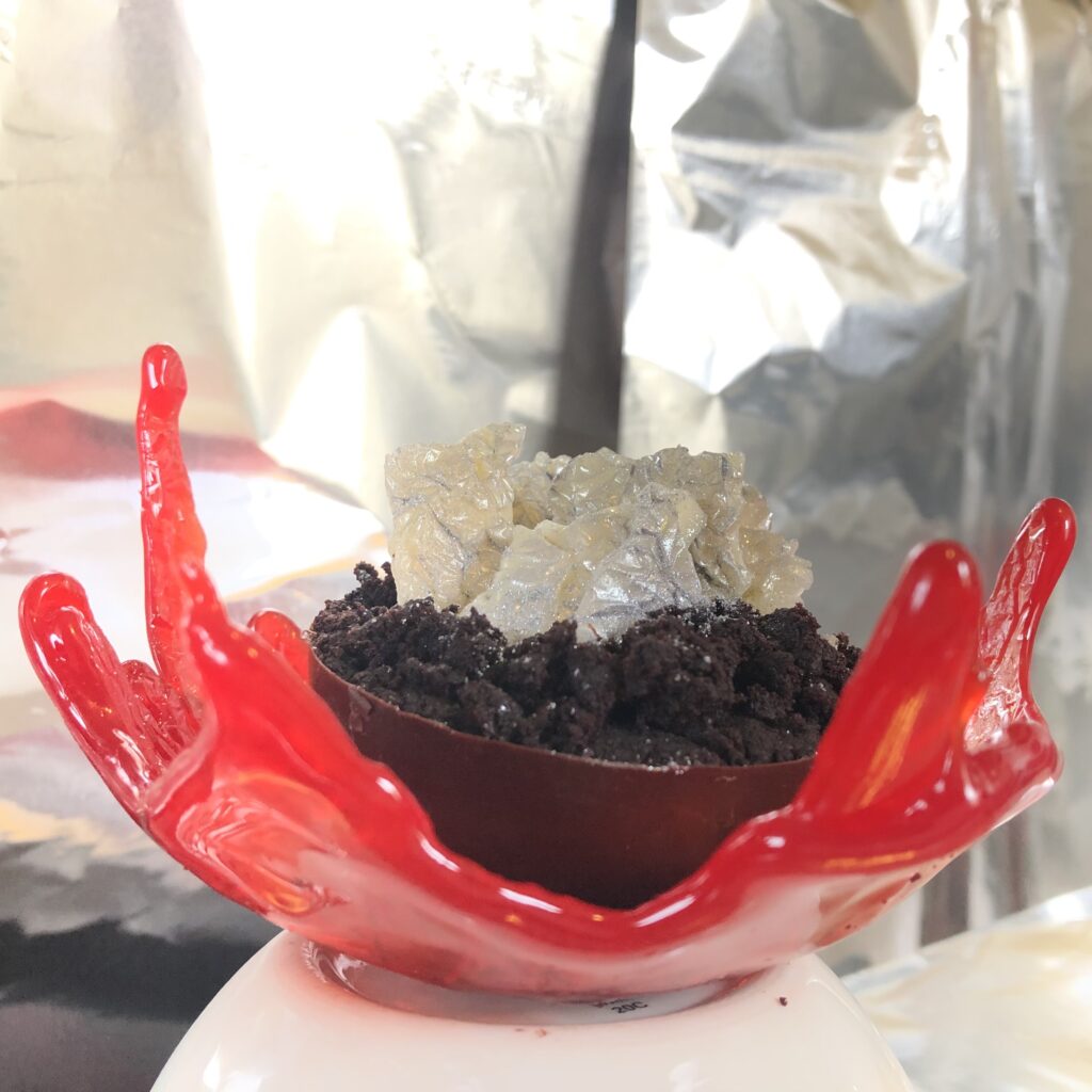 Crackled, textured white chocolate shards with shades of grey and silver stand at various angles in the center of a chocolate cake-filled chocolate hemisphere. The hemisphere is held by a red sugar sail sitting concave up on an upside down ceramic bowl with a silver background.