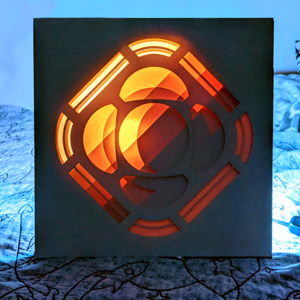 A paper lightbox of the Psyche logo that is divided into multiple layers, which indivudually appear to be abstract shapes. When viewed from the front, the layers line up to depict the Psyche logo. The lighting causes the box to go from a lighter shade in the top left to a darker shade in the bottom right.