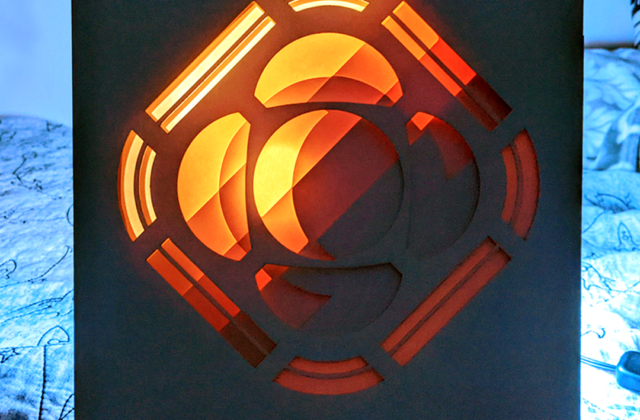 A paper lightbox of the Psyche logo that is divided into multiple layers, which indivudually appear to be abstract shapes. When viewed from the front, the layers line up to depict the Psyche logo. The lighting causes the box to go from a lighter shade in the top left to a darker shade in the bottom right.