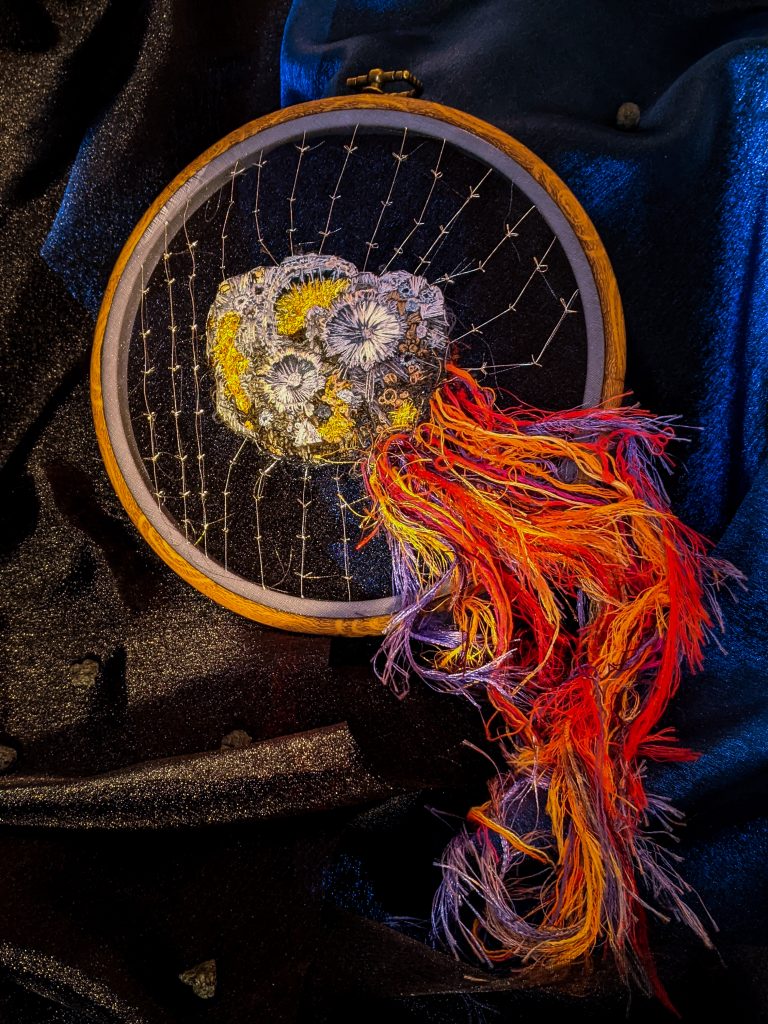 The Psyche asteroid embroidered with silver stitching to represent magnetic fields. A trail follows behind Psyche and off of the embroidery wheel.