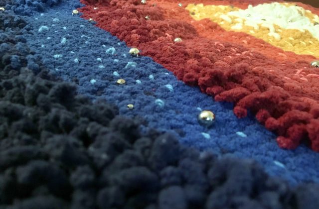 A close-up view of a tapestry of woven yarn in dark blue, light blue, red, light red, orange, yellow, and cream.