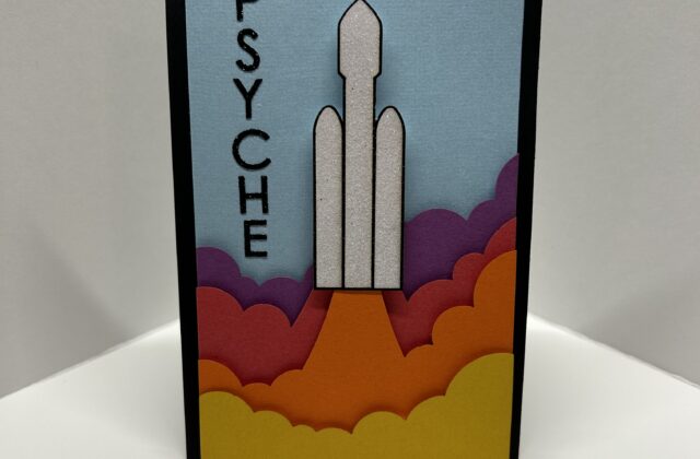 The front of the card shows a Falcon-9 rocket launch with layers of exhaust clouds colored to match the Psyche logo. The word “PSYCHE” is written down the side. The card is opened and the Psyche spacecraft unfolds and pops out of the middle. Behind the spacecraft layered paper recreates the rough and cratered surface of the asteroid.