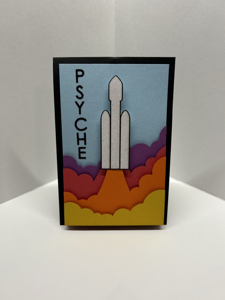 The front of the card shows a Falcon-9 rocket launch with layers of exhaust clouds colored to match the Psyche logo. The word “PSYCHE” is written down the side. The card is opened and the Psyche spacecraft unfolds and pops out of the middle. Behind the spacecraft layered paper recreates the rough and cratered surface of the asteroid.