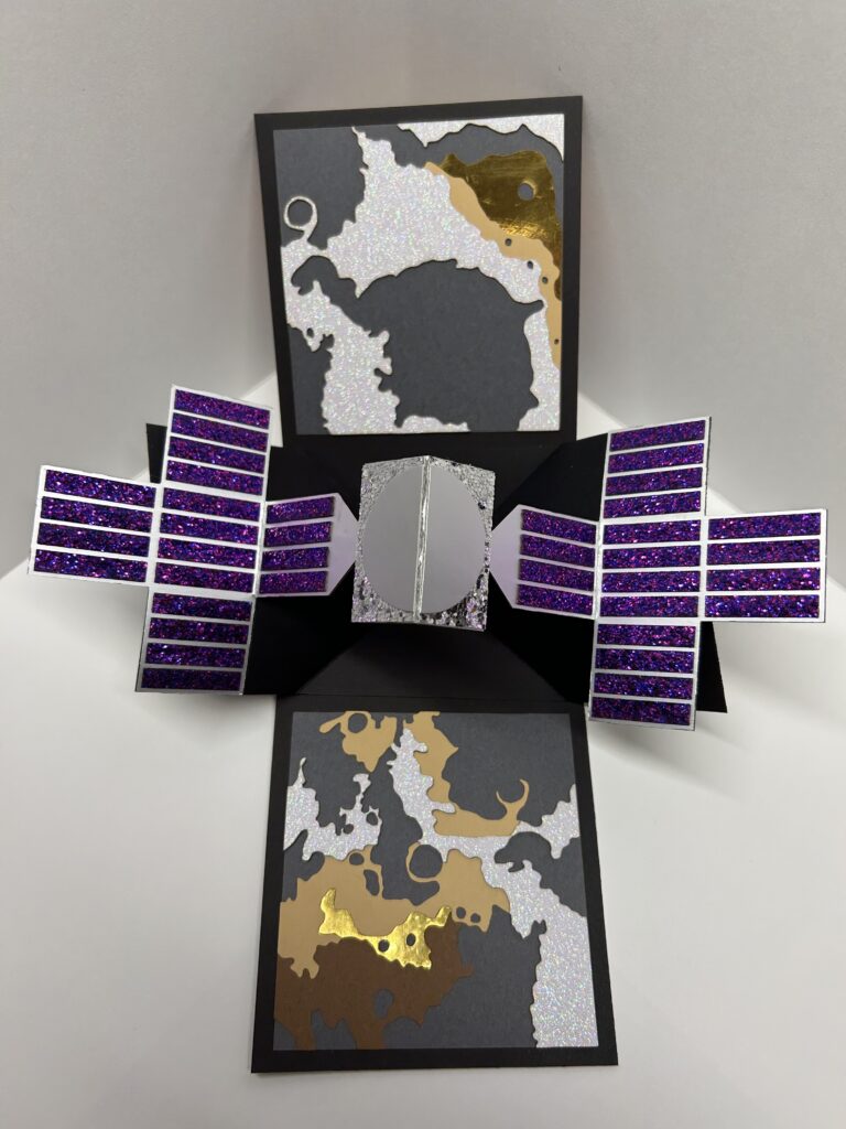 The front of the card shows a Falcon-9 rocket launch with layers of exhaust clouds colored to match the Psyche logo. The word “PSYCHE” is written down the side. The card is opened and the Psyche spacecraft unfolds and pops out of the middle. Behind the spacecraft layered paper recreates the rough and cratered surface of the asteroid.