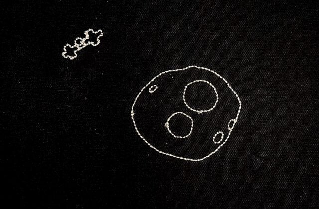 An embroidered stop-motion animation depicting the asteroid Psyche rotating in 3D. As it spins, a twinkle of light reveals the Psyche spacecraft, which flies closer until only one cross-shaped solar panel is visible. The scene then morphs into the colorful Psyche mission logo.