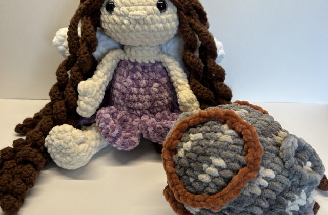 The doll is seated. She has pale skin and wears a variegated purple dress. The skirt of the dress has the shape of flower petals. She has long, curly brown hair that cascades down to the floor, and large white wings peak out from behind it. The asteroid is an oblong shape and an overall gray color with spots of white and brown. It has embroidered craters which protrude from the surface.