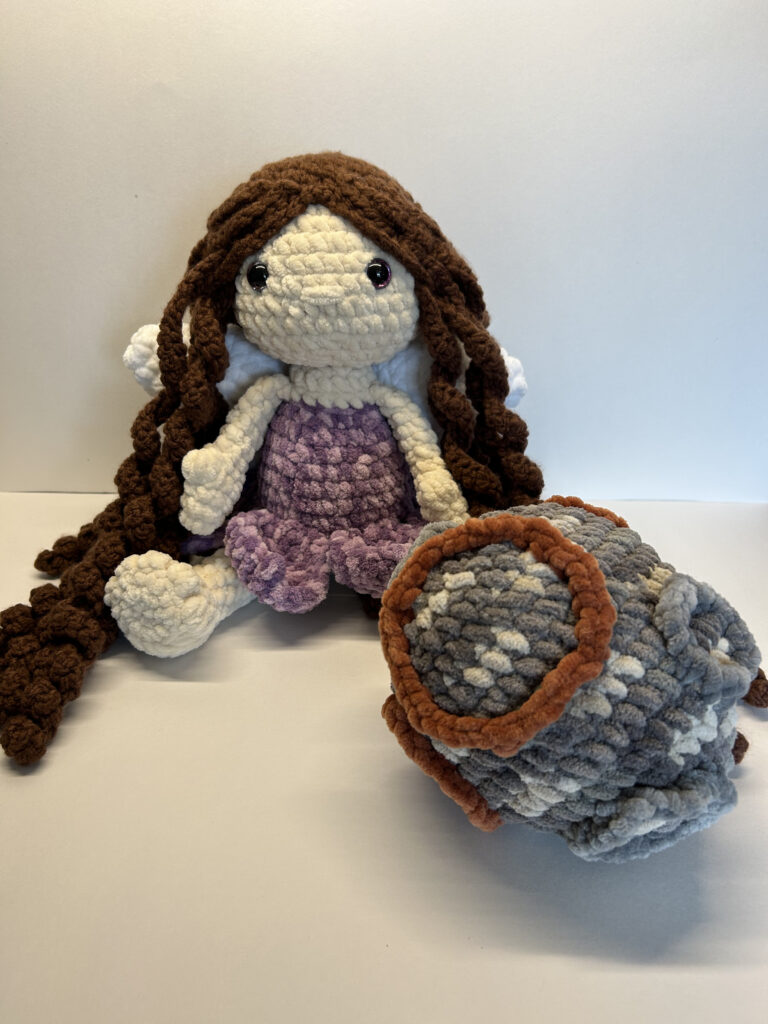 The doll is seated. She has pale skin and wears a variegated purple dress. The skirt of the dress has the shape of flower petals. She has long, curly brown hair that cascades down to the floor, and large white wings peak out from behind it. The asteroid is an oblong shape and an overall gray color with spots of white and brown. It has embroidered craters which protrude from the surface.