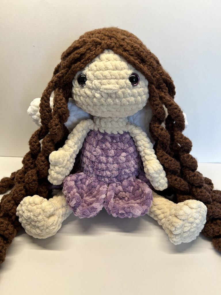 The doll is seated. She has pale skin and wears a variegated purple dress. The skirt of the dress has the shape of flower petals. She has long, curly brown hair that cascades down to the floor, and large white wings peak out from behind it. The asteroid is an oblong shape and an overall gray color with spots of white and brown. It has embroidered craters which protrude from the surface.