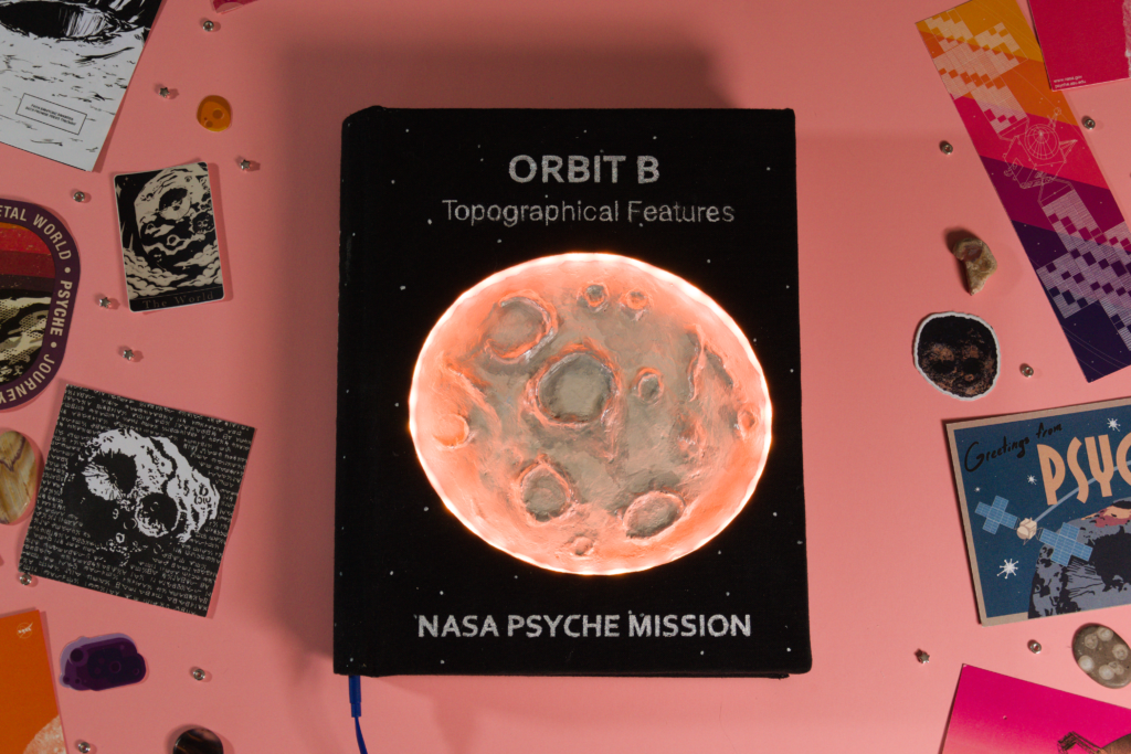 This book is the second in a series about the four orbits the spacecraft will inhabit once it reaches Psyche. The cover features a clay interpretation of the asteroid surrounded by a ring of lights mimicking day and night. The back blurb talks about the mission of orbit B. The interior is hand sewn and finished with marbled end pages.