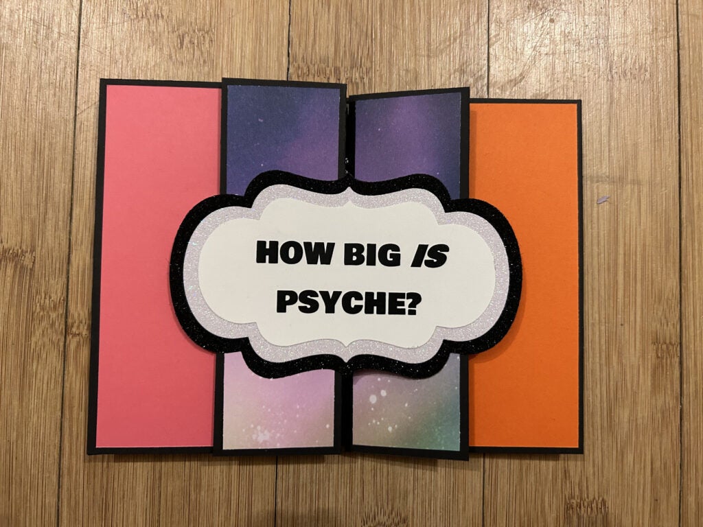 The front of the card asks the question “How big is Psyche?”. When the card unfolds, it expands to three times its original size. Colored panels in the middle make up the Psyche spacecraft. Two larger panels on either side give facts about Psyche’s size and weight, stating that the spacecraft weighs as much as four cows, and is the size of a tennis court with its solar arrays extended.