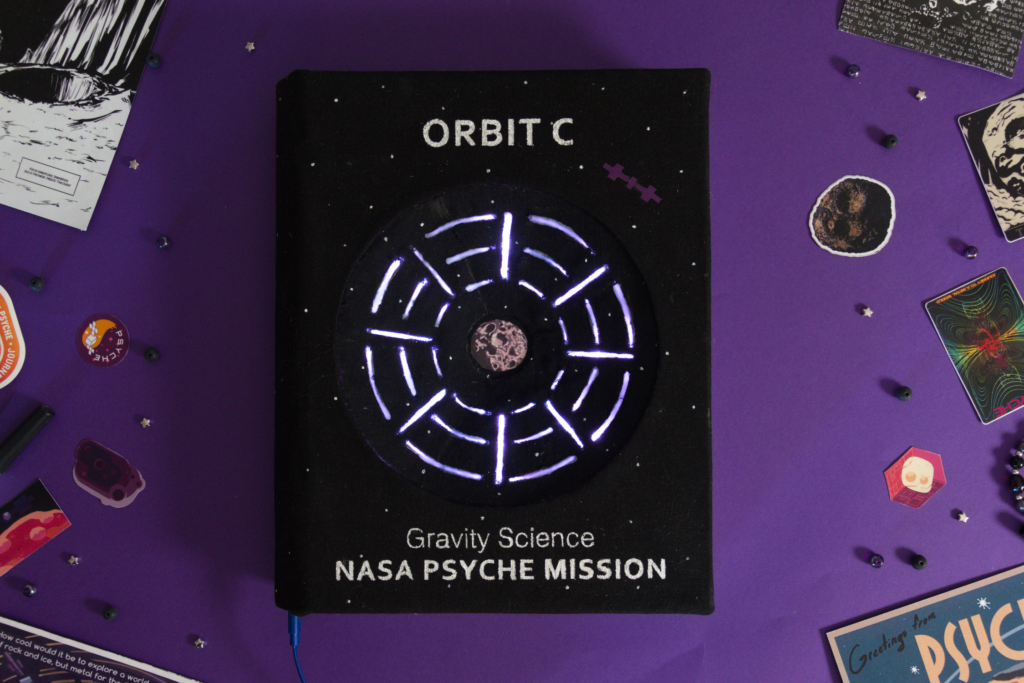 Orbit C Notebook is the third in the series discussing the gravity field science. The cover features an inset cone, mimicking space time, and LED’s with an animation of direction of acceleration. The interior is hand sewn with circles cut out of the pages to allow the cone to sit flush inside and finished with marbled end pages.